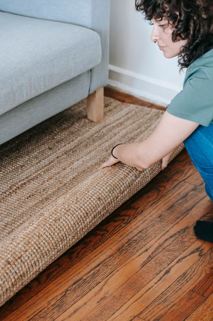 A woman with curly dark hair, wearing a teal shirt and dark pants, is carefully lifting and adjusting a woven brown rug on a polished wooden floor in a living room. The room features a light grey upho